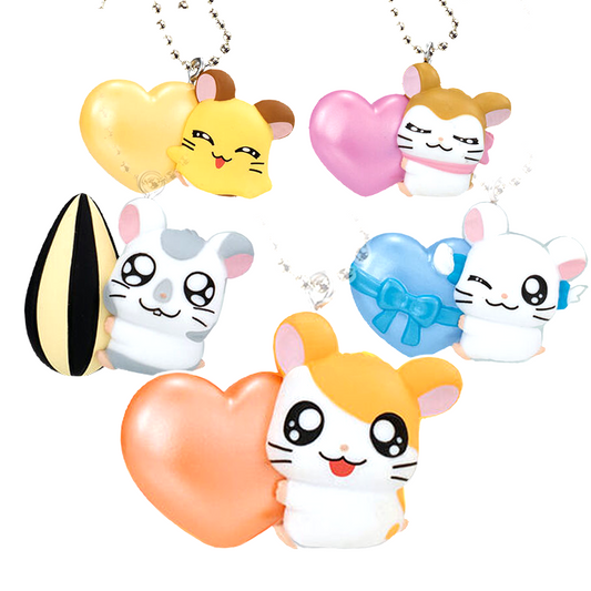 Immagine promozionale di 5 ciondoli portachiavi gashapon della serie Hamtaro disposti su sfondo bianco. Al centro in basso spicca il criceto Hamtaro (bianco e arancione) sorridente mentre abbraccia un grande cuore arancione perlato. Attorno a lui, partendo da sinistra, troviamo: Ghiotto (bianco e grigio chiaro) aggrappato a un enorme seme di girasole a strisce nere e gialle; in alto a sinistra la piccola Penelope (gialla e marrone) dietro a un cuore giallo.