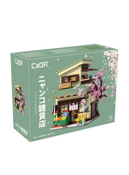 Scatola e modello assemblato del set di costruzioni CaDA "Kitty's Grocery" (C66015W). Raffigura un tipico negozietto giapponese a due piani con tetto verde, un grande albero di ciliegio rosa fiorito sul lato, espositori esterni con riviste, un gatto arancione e l'interno del negozio visibile grazie a una parete apribile. Sulla scatola è indicata la presenza di 921 pezzi e del sistema LED.