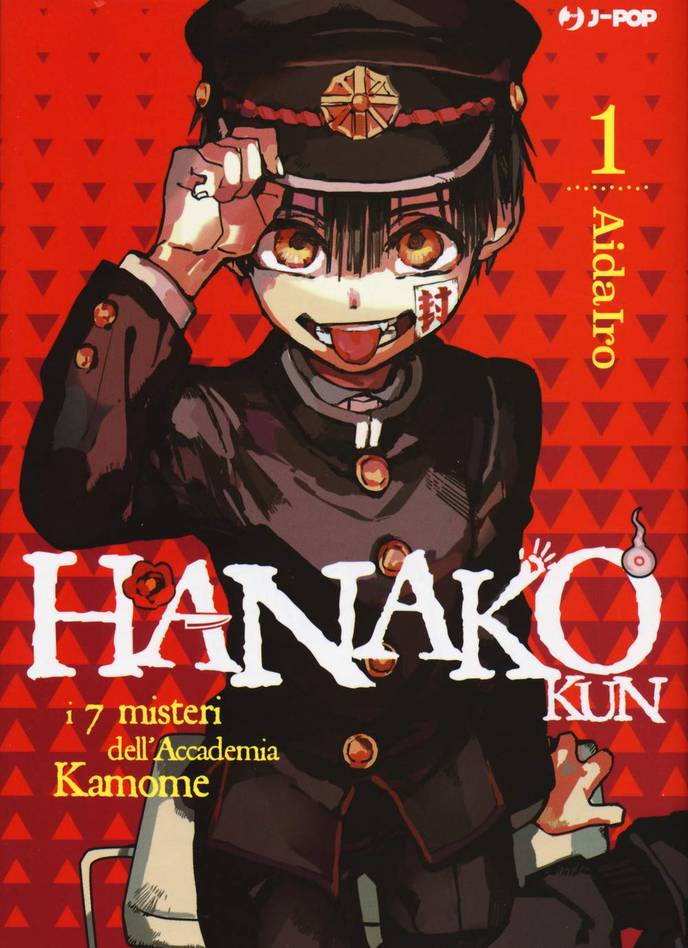 Copertina del manga Hanako-kun e i sette misteri dell'Accademia Kamome Vol. 1, edito da J-POP Manga. L'illustrazione su uno sfondo rosso acceso con un pattern a triangoli scuri mostra in primo piano il giovane Hanako. Il ragazzo indossa una vecchia divisa scolastica nera con bottoni dorati e un berretto abbinato. Ha grandi occhi ambrati, fa una linguaccia impertinente e presenta un sigillo di carta bianca con scritte rosse incollato sulla guancia sinistra. Con la mano destra si tocca la tesa del cappello.