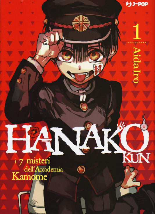 Copertina del manga Hanako-kun e i sette misteri dell'Accademia Kamome Vol. 1, edito da J-POP Manga. L'illustrazione su uno sfondo rosso acceso con un pattern a triangoli scuri mostra in primo piano il giovane Hanako. Il ragazzo indossa una vecchia divisa scolastica nera con bottoni dorati e un berretto abbinato. Ha grandi occhi ambrati, fa una linguaccia impertinente e presenta un sigillo di carta bianca con scritte rosse incollato sulla guancia sinistra. Con la mano destra si tocca la tesa del cappello.
