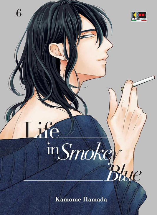 Copertina del manga Life in Smokey Blue Vol. 6. Illustrazione elegante che ritrae di profilo un ragazzo dai lunghi capelli scuri. Indossa un maglione blu scuro scivolato su una spalla, tenendo con delicatezza una sigaretta accesa nella mano destra. Lo sfondo è di un colore grigio chiaro e neutro, con il titolo dell'opera scritto al centro e il nome dell'autrice, Kamome Hamada, in basso.