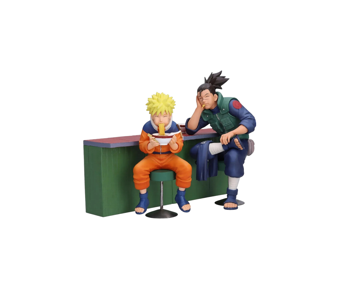 Set di due figure da collezione Banpresto raffiguranti Naruto Uzumaki e Iruka Umino seduti al bancone del chiosco Ichiraku Ramen. Naruto è seduto su uno sgabello verde mentre mangia con gioia una ciotola di ramen con le bacchette, affiancato da Iruka Umino che lo guarda con un sorriso affettuoso appoggiando la testa sulla mano.