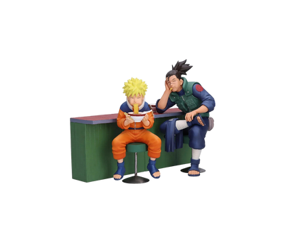Set di due figure da collezione Banpresto raffiguranti Naruto Uzumaki e Iruka Umino seduti al bancone del chiosco Ichiraku Ramen. Naruto è seduto su uno sgabello verde mentre mangia con gioia una ciotola di ramen con le bacchette, affiancato da Iruka Umino che lo guarda con un sorriso affettuoso appoggiando la testa sulla mano.