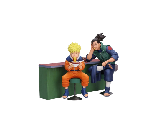 Set di due figure da collezione Banpresto raffiguranti Naruto Uzumaki e Iruka Umino seduti al bancone del chiosco Ichiraku Ramen. Naruto è seduto su uno sgabello verde mentre mangia con gioia una ciotola di ramen con le bacchette, affiancato da Iruka Umino che lo guarda con un sorriso affettuoso appoggiando la testa sulla mano.