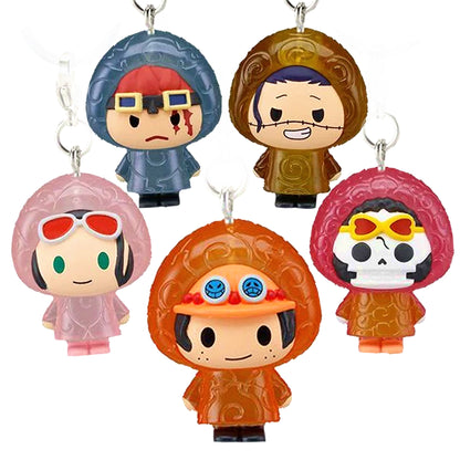 Immagine promozionale di 5 ciondoli gashapon della serie One Piece disposti su uno sfondo bianco pulito. I personaggi, in stile super deformed (chibi), indossano degli impermeabili traslucidi decorati con il classico motivo a spirale dei Frutti del Diavolo. Al centro in primo piano spicca Portgas D. Ace con il suo iconico cappello e un impermeabile arancione. In alto a sinistra si trova Eustass Kid con occhialoni, cicatrice sul viso e impermeabile blu-grigio scuro.